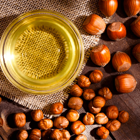 Be Bohemian Botanical Sea Serum Hazelnut Oil. A top down view onto a round clear container with bright yellow clear oil sits on top of a woven place mat next to an assortment of hazelnuts.