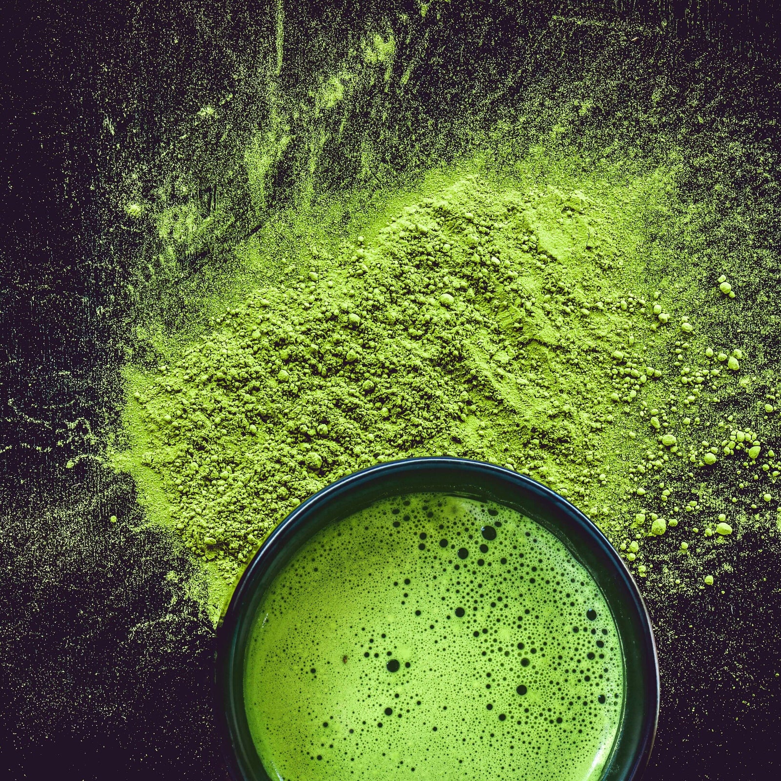 Be Bohemian Botanical Sea Serum Organic Green Tea. A dark cup of hot green liquid with black bubbles in it sits in a little mound of brilliant green powder messily spread on a black countertop or table.