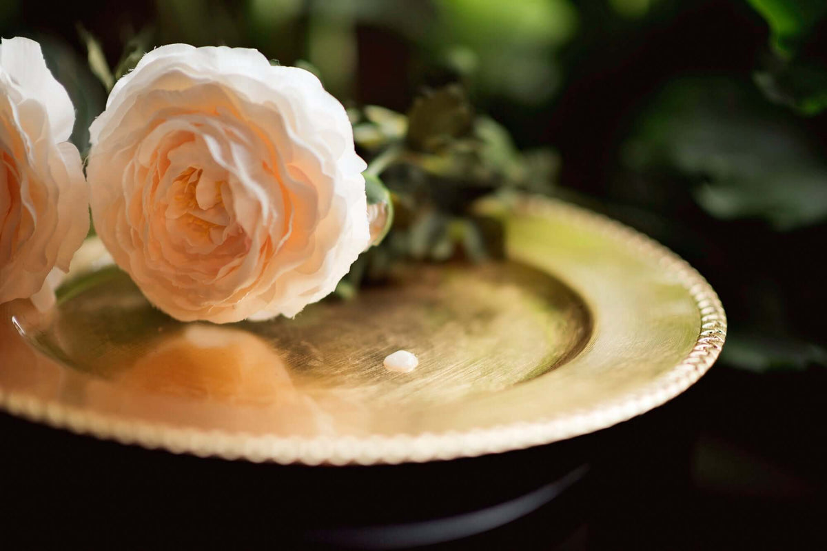 A drop of Be Bohemian Botanical Sea Serum on a golden platter next to a pink rose with a dark background.